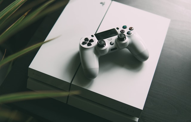 Playstation 4 ontop of a table with a controller on top of the playstation, and slightly blocked by a plant on the left side of the image.
